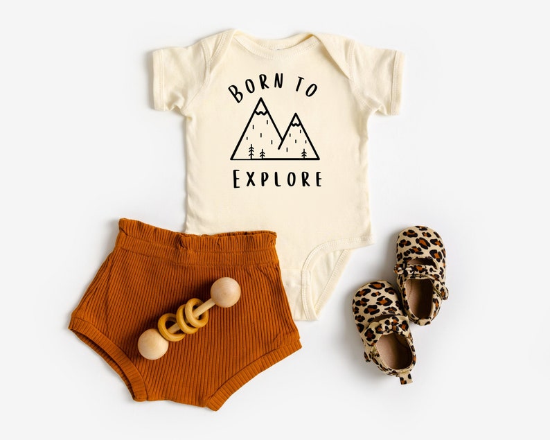 May include: A cream colored baby onesie with the text "Born to Explore" and a black mountain graphic. The onesie is paired with brown corduroy shorts and leopard print baby shoes.