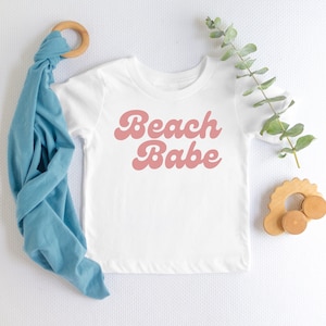 May include: A white baby onesie with pink text that reads "Beach Babe" in a retro font style. The onesie is laid flat on a white surface with a blue fabric swaddle and a wooden toy nearby.