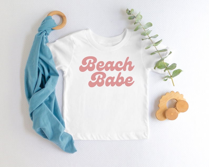 May include: A white baby onesie with pink text that reads "Beach Babe" in a retro font style. The onesie is laid flat on a white surface with a blue fabric swaddle and a wooden toy nearby.