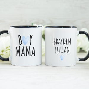 Puede incluir: Dos tazas de cer&aacute;mica blanca con asas negras y bordes negros. Una taza dice "Boy Mama" con un coraz&oacute;n azul. La otra taza dice "Brayden Julian" con un coraz&oacute;n azul.