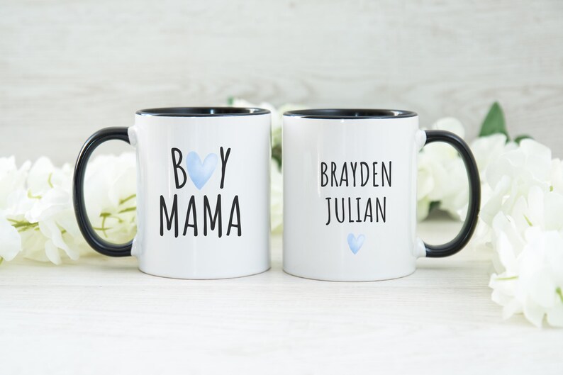 Puede incluir: Dos tazas de cer&aacute;mica blanca con asas negras y bordes negros. Una taza dice "Boy Mama" con un coraz&oacute;n azul. La otra taza dice "Brayden Julian" con un coraz&oacute;n azul.