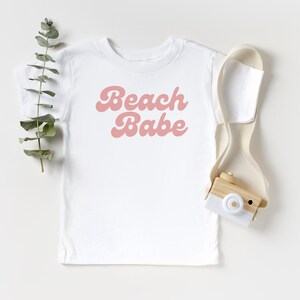 May include: White t-shirt with pink retro font that says "Beach Babe". A small wooden toy camera with a white strap is on the right side of the image.