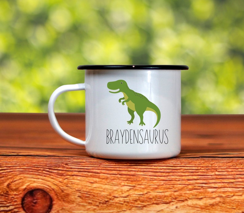 K&ouml;nnte beinhalten: Wei&szlig;er Emaillebecher mit schwarzem Rand und Henkel. Auf der Vorderseite befindet sich eine gr&uuml;ne Dinosaurier-Grafik mit dem Wort "BRAYDENSAURUS" in schwarzer Schrift darunter. Der Becher steht auf einer Holzoberfl&auml;che.