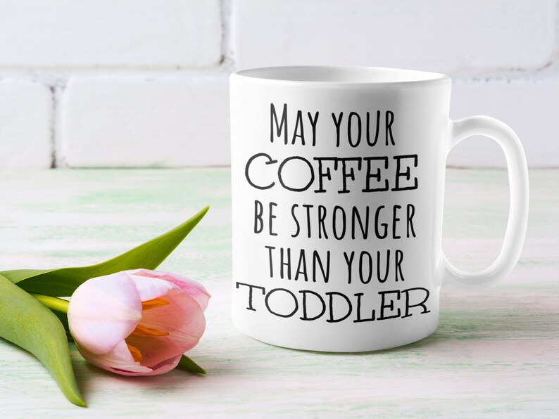Pode incluir: Caneca de caf&eacute; de cer&acirc;mica branca com o texto "MAY YOUR COFFEE BE STRONGER THAN YOUR TODDLER" em preto. Uma tulipa rosa com folhas verdes est&aacute; em primeiro plano. O fundo &eacute; uma parede de tijolos brancos e uma superf&iacute;cie verde claro.