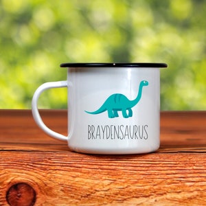 K&ouml;nnte beinhalten: Wei&szlig;er Emaillebecher mit schwarzem Rand und Henkel. Er zeigt eine t&uuml;rkisfarbene Dinosaurier-Illustration und das Wort "BRAYDENSAURUS" in schwarzem Text. Der Becher steht vor einem verschwommenen gr&uuml;nen Hintergrund.