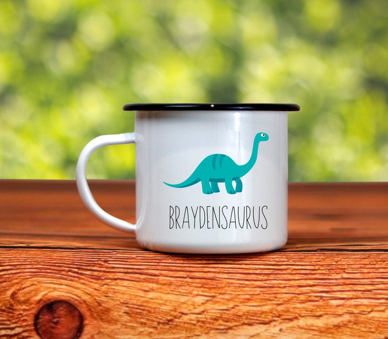 K&ouml;nnte beinhalten: Wei&szlig;er Emaillebecher mit schwarzem Rand und Henkel. Er zeigt eine t&uuml;rkisfarbene Dinosaurier-Illustration und das Wort "BRAYDENSAURUS" in schwarzem Text. Der Becher steht vor einem verschwommenen gr&uuml;nen Hintergrund.