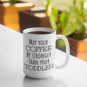 Pode incluir: Caneca de caf&eacute; de cer&acirc;mica branca com o texto "May your coffee be stronger than your toddler" em preto. A caneca est&aacute; cheia de caf&eacute; escuro e tem uma al&ccedil;a curva. A caneca est&aacute; sobre uma superf&iacute;cie branca.