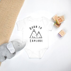May include: White baby onesie with black text that reads "Born to Explore" and a graphic of two mountains with trees and rain.