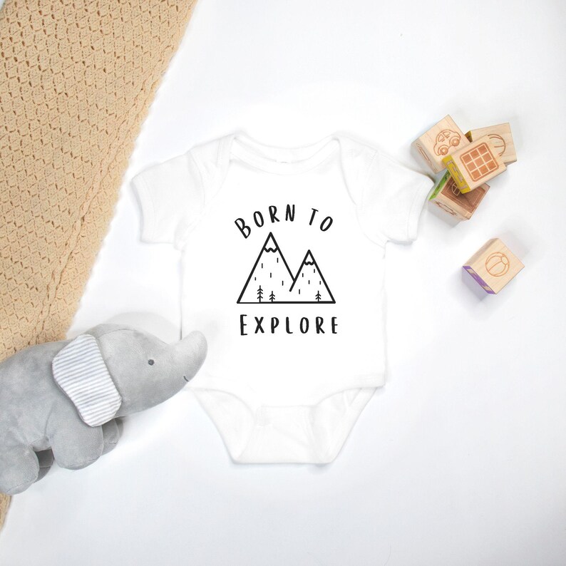 May include: White baby onesie with black text that reads "Born to Explore" and a graphic of two mountains with trees and rain.