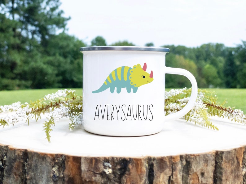K&ouml;nnte beinhalten: Wei&szlig;er Emaillebecher mit silbernem Rand und wei&szlig;em Henkel. Der Becher zeigt einen Cartoon-Triceratops in Blau und Gelb mit dem Namen "Averysaurus" darunter in Schwarz.