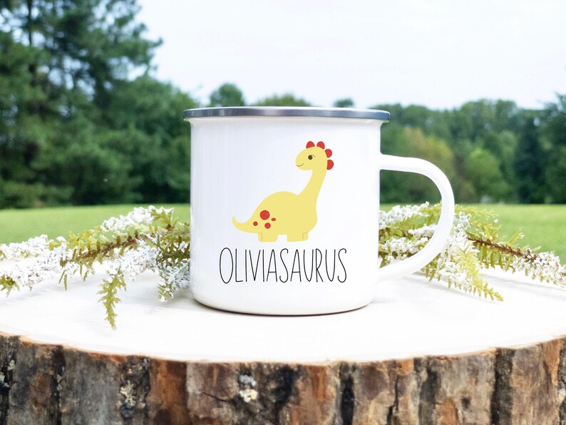 K&ouml;nnte beinhalten: Wei&szlig;er Emaillebecher mit silbernem Rand und Henkel. Der Becher zeigt eine gelbe Dinosaurier-Illustration mit roten Punkten und einem roten Kamm. Der Text "OLIVIASAURUS" ist darunter in Schwarz gedruckt.