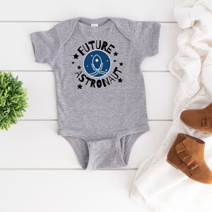 May include: Gray baby bodysuit with a blue and black rocket graphic and the text "Future Astronaut" in a circle.