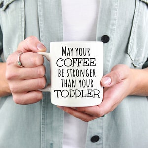 Pode incluir: Uma caneca de caf&eacute; de cer&acirc;mica branca com o texto "MAY YOUR COFFEE BE STRONGER THAN YOUR TODDLER" em letras pretas. A caneca &eacute; segurada por uma pessoa que usa uma jaqueta verde claro.