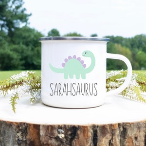 K&ouml;nnte beinhalten: Wei&szlig;er Emaillebecher mit silbernem Rand und Henkel. Der Becher zeigt eine hellgr&uuml;ne Dinosaurier-Illustration mit lila Stacheln und das Wort "SARAHSAURUS" in schwarzer Schrift. Der Becher steht auf einer Holzoberfl&auml;che mit Gr&uuml;n.