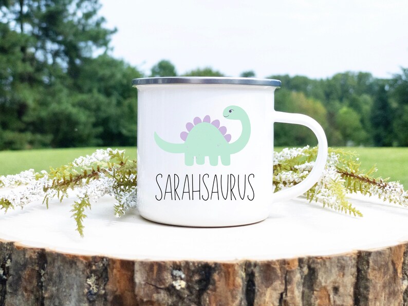 K&ouml;nnte beinhalten: Wei&szlig;er Emaillebecher mit silbernem Rand und Henkel. Der Becher zeigt eine hellgr&uuml;ne Dinosaurier-Illustration mit lila Stacheln und das Wort "SARAHSAURUS" in schwarzer Schrift. Der Becher steht auf einer Holzoberfl&auml;che mit Gr&uuml;n.