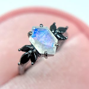 May include: A close-up of a ring with a large, iridescent, hexagonal gemstone as the focal point. The gemstone is surrounded by smaller, black, teardrop-shaped stones and set on a dark metal band. The ring is displayed against a soft pink background.