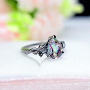 May include: A black metal ring with a pear-shaped, iridescent gemstone. The ring features a leaf and vine design on the band and around the gemstone. Small black stones accent the setting. The gemstone displays a range of colors, including green, purple, and blue.