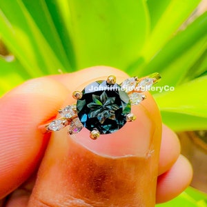 May include: A gold ring with a large, dark blue gemstone surrounded by smaller, clear gemstones. The ring is set with a floral design and the text "JohnfinejewelryCo" is visible on the band.