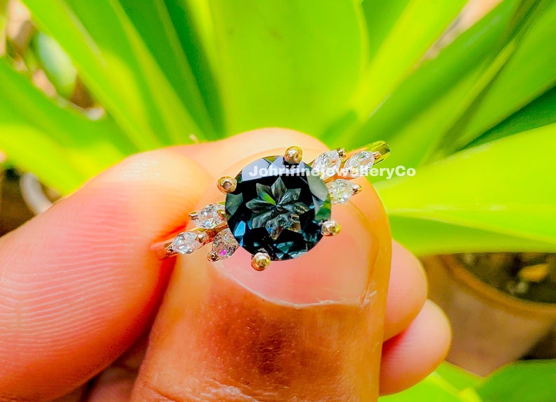May include: A gold ring with a large, dark blue gemstone surrounded by smaller, clear gemstones. The ring is set with a floral design and the text "JohnfinejewelryCo" is visible on the band.