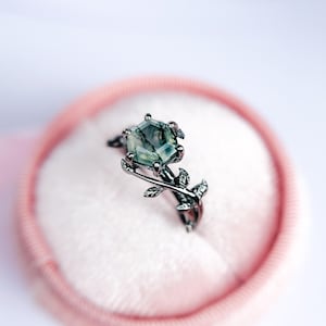 Puede incluir: Un anillo de plata con una piedra preciosa de ágata verde engastada en una forma hexagonal. El anillo presenta un delicado diseño de hojas que se envuelve alrededor de la banda.