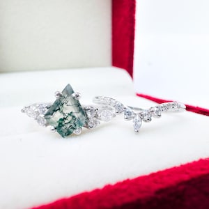 May include: A silver ring set with a green moss agate gemstone and a matching silver band with small clear stones. The rings are displayed in a red velvet box.