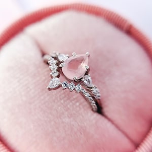 Op de afbeelding: Een delicate ringset met een peervormige roze edelsteen omringd door kleinere heldere edelstenen. De ring bevindt zich in een roze fluwelen ringdoos.
