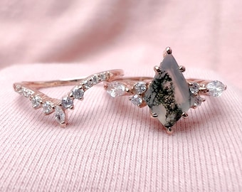 May include: A pair of rose gold rings with a pear-shaped moss agate gemstone and small clear stones. The rings are on a pink fabric background.