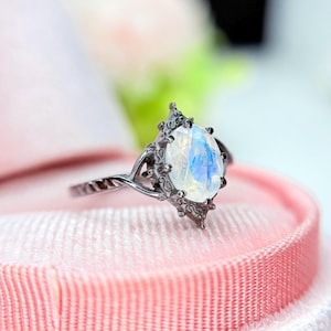 May include: A silver ring with a pear-shaped, iridescent gemstone. The ring has an ornate, dark metal setting with a textured band. The gemstone displays a play of blue and white colors, set against a pink background.