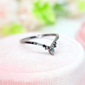 May include: A delicate, black-toned ring with a chevron design. The ring features a central, light-colored gemstone and is accented with small, black stones. The ring is set on a soft, pink surface, creating a contrast of colors and textures.