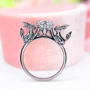 May include: A silver ring with a large, clear gemstone at the center, flanked by two stylized figures. The ring's design features intricate details and a polished finish, set against a soft pink background.