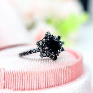 May include: A black gemstone ring with a starburst design. The ring features a large, faceted black stone at the centre, surrounded by smaller black stones. The band is also adorned with black stones. The ring is set on a pink surface.
