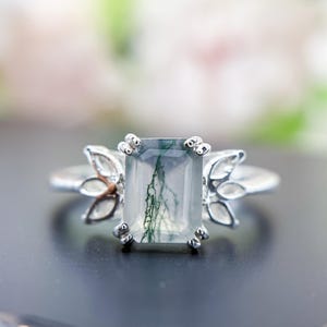 May include: A silver ring featuring an emerald-cut gemstone with green inclusions. The ring has a floral design on the sides and is set against a blurred background. The gemstone is approximately 0.5 inches in length.
