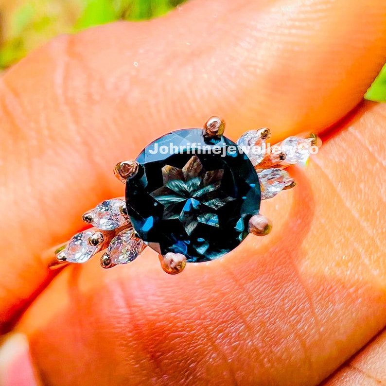 May include: A silver ring with a large, round, dark blue gemstone surrounded by smaller, clear gemstones. The text "Johrifinejewellersco" is visible on the ring.
