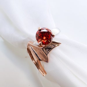 May include: A rose gold ring with a red gemstone set in a prong setting. The ring band is shaped like a stylized wing.