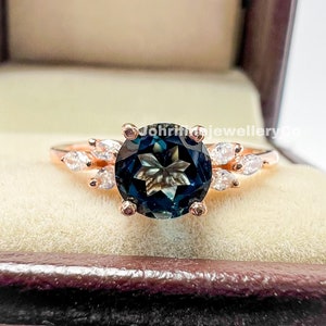 May include: A rose gold ring with a large, round, dark blue gemstone surrounded by smaller, clear gemstones. The ring is in a velvet ring box.