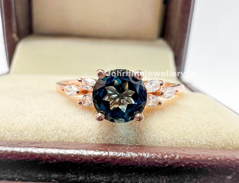 May include: A rose gold ring with a large, round, dark blue gemstone surrounded by smaller, clear gemstones. The ring is in a velvet ring box.