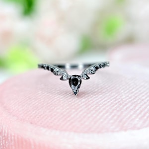 Könnte beinhalten: Ein silberfarbener Ring mit einem birnenförmigen schwarzen Edelstein in der Mitte. Der Ring hat ein geschwungenes Design mit kleineren schwarzen Steinen entlang des Bandes, was ein zartes und elegantes Aussehen erzeugt. Der Ring liegt auf einer rosa Oberfläche.