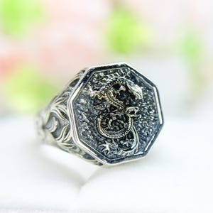 May include: A silver ring with an octagonal face featuring a detailed dragon design. The ring has an ornate, textured band with a floral pattern. The dragon is depicted in a coiled pose, with intricate scales and features.