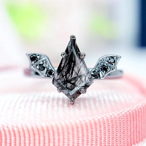 Könnte beinhalten: Silberring mit einem großen, facettierten, schwarzen und klaren Edelstein in einer einzigartigen Form. Der Ring hat kleine, schwarze Edelsteine und flügelförmige Akzente. Präsentiert auf rosa Oberfläche.