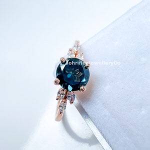 May include: A rose gold ring with a large, round, dark blue gemstone surrounded by smaller, clear gemstones. The ring is set on a white background.
