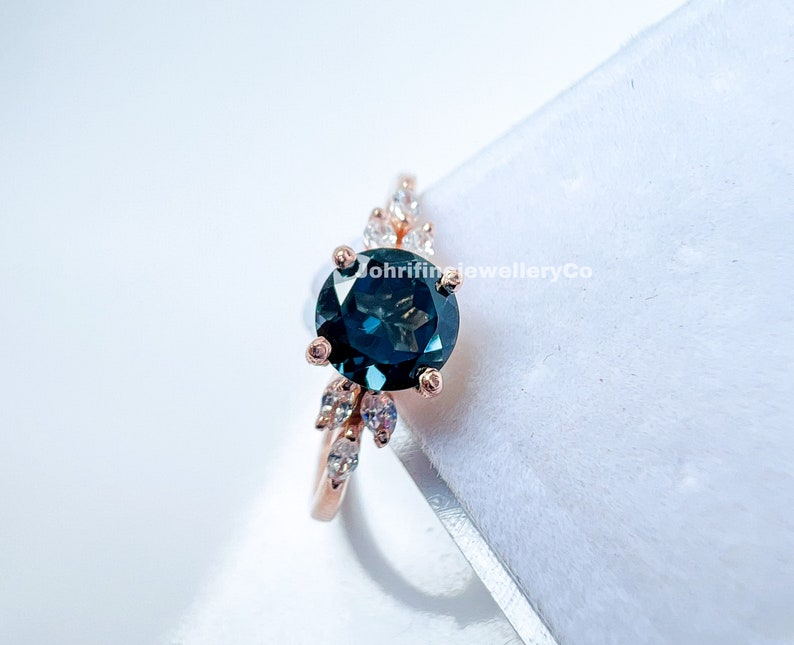 May include: A rose gold ring with a large, round, dark blue gemstone surrounded by smaller, clear gemstones. The ring is set on a white background.