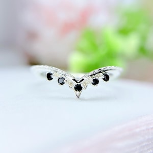 Może przedstawiać: Srebrny pierścionek o kształcie litery V. Pierścionek zdobiony jest małymi, okrągłymi czarnymi kamieniami szlachetnymi. Wzór pierścionka jest delikatny i skomplikowany, z naciskiem na kontrast między srebrnym metalem a czarnymi kamieniami.