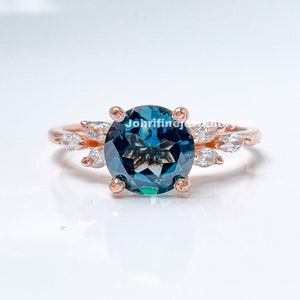 May include: A rose gold ring with a large, round, teal blue gemstone surrounded by smaller, clear gemstones. The ring has a delicate, leaf-like design.