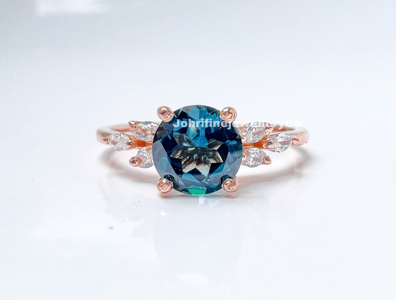 May include: A rose gold ring with a large, round, teal blue gemstone surrounded by smaller, clear gemstones. The ring has a delicate, leaf-like design.