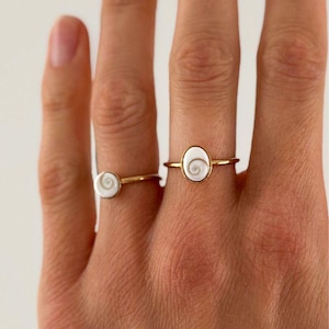 Puede incluir: Dos anillos dorados con diseños blancos en forma de espiral. Los anillos están en una mano, mostrando la delicada joyería. Los anillos tienen una fina banda dorada y un centro blanco de forma ovalada con un diseño en espiral.