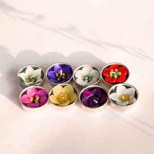 Könnte beinhalten: Acht kleine, runde, weiße Votivkerzenhalter mit floralen Designs in verschiedenen Farben, darunter Lila, Rosa, Gelb, Rot und Weiß. Die Kerzen sind in einer Reihe auf einer weißen Oberfläche angeordnet.