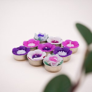 Könnte beinhalten: Eine Ansammlung von blütenförmigen Kerzen in verschiedenen Farben, darunter Lila, Rosa und Weiß. Jede Kerze befindet sich in einem silbernen Metallbehälter. Die Kerzen sind in einer Gruppe angeordnet und bilden eine dekorative Präsentation.