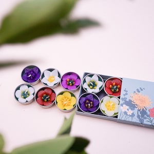 Puede incluir: Un conjunto de velas con forma de flor en varios colores, incluyendo morado, blanco, rojo y amarillo, dispuestas en latas plateadas. Las velas se exhiben en una caja azul claro con ilustraciones florales. El tema general es decorativo y floral.