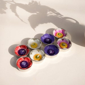Könnte beinhalten: Sortiment von acht Teelichtern in verschiedenen Farben und floralen Designs. Die Teelichter sind in einem Kreis auf einer weißen Oberfläche angeordnet.