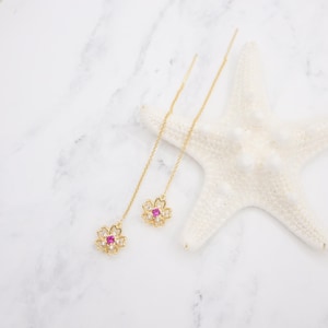 May include: A pair of gold earrings with delicate chains and pink flower charms. Each flower charm has a pink gemstone in the center and smaller clear gemstones around it.